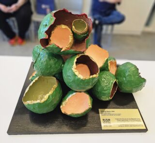 Tightly framed photograph of green, gold, and peach painted bulbous sculpture sitting on a table top. Not in focus, there are a few people sitting in chairs in the foreground (facing the artwork).