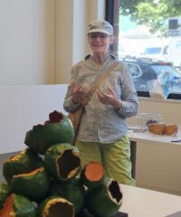 A woman in a blue button down shirt, green pants, and a newsies cap, standing in the foreground speaking and gesturing with her hands towards the partially visible bulbous green and orange sculpture in the foreground sitting on a table top
