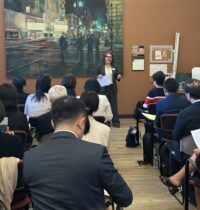 Woman in a business suit, speaking and holding documents, standing at the front of a room of individuals seated looking towards her.