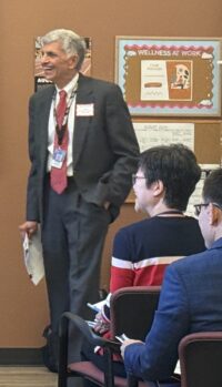 Man in a business suit, holding papers in his hand by his side, standing at the front of a room with people seated in chairs looking in his direction, looking slightly to his right smiling and laughing in response to something