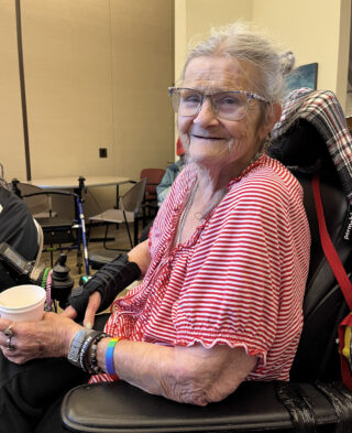 An elderly white presenting woman with glasses in a red and white striped shirt sits in a mobility chair holding a paper cup, she is smiling and turning her face towards the camera