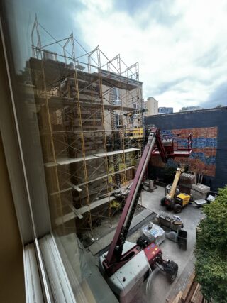 A photo taken from inside shows an active construction space in courtyard. Two large machines sit in the courtyard with lifting arms. To the left is a multi-story scaffolding covering the whole side of the building.