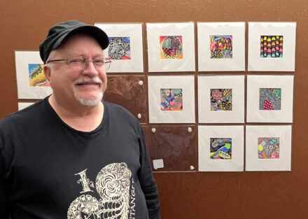 An older white man with glasses, a white close-trimmed beard, and a black brimmed hat, smiles into the camera standing in front of a collection of square abstract, detailed illustrations hanging on the wall behind him.