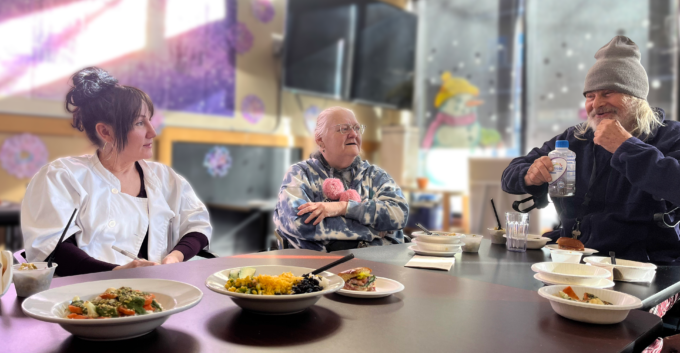Three adults sit around a long table, with multiple dishes of food in front of them, the man at the end of the table to the right is smiling mid-sentence, with his hand on this chin, while the woman next to him looks on with her arms crossed, and the woman at the other edge of the table also looks on while wearing chef's whites.