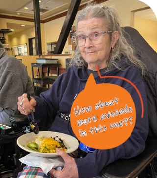 Older white woman sitting in a mobility device holds a plate of food and looks at the camera. An orange cartoon speech bubble has text on it reading "how about more avocado in this one??"