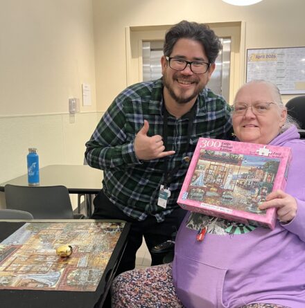 To the side of a completed puzzle on a table, sits an older white woman holding a puzzle box to the camera and smiling, standing between her and the puzzle table is a younger gentleman with glasses who is leaning forward slightly, smiling at the camera