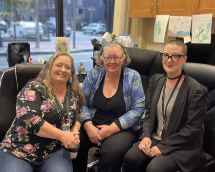Three white women sitting in rolling office chairs, close together smiling up at the camera