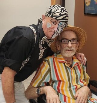 White man wearing a black & white bandana on their head and half a face of black & white face paint and jewels leans with one arm around the shoulders of an older white man seated in a wheelchair, wearing brightly colored striped ensemble