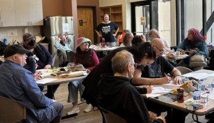 Several tables of full of people are sitting and smiling and talking in a room together, everyone has papers and pens in front of them. They are participating in a quarterly Member Forum with Maybelle Center's Neighborhood Programs in Portland, Oregon. During these meetings our community provides vital feedback and input to help shape all programs, in this meeting a thorough surveying of input for the community room mural is underway.