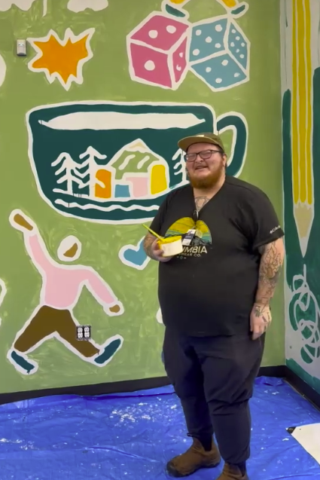White individual with a bread and glasses stands holding a cup with paint and a paintbrush, smiling to the camera and standing in front of the Community Room mural in-process at Maybelle Center in Portland, OR. 