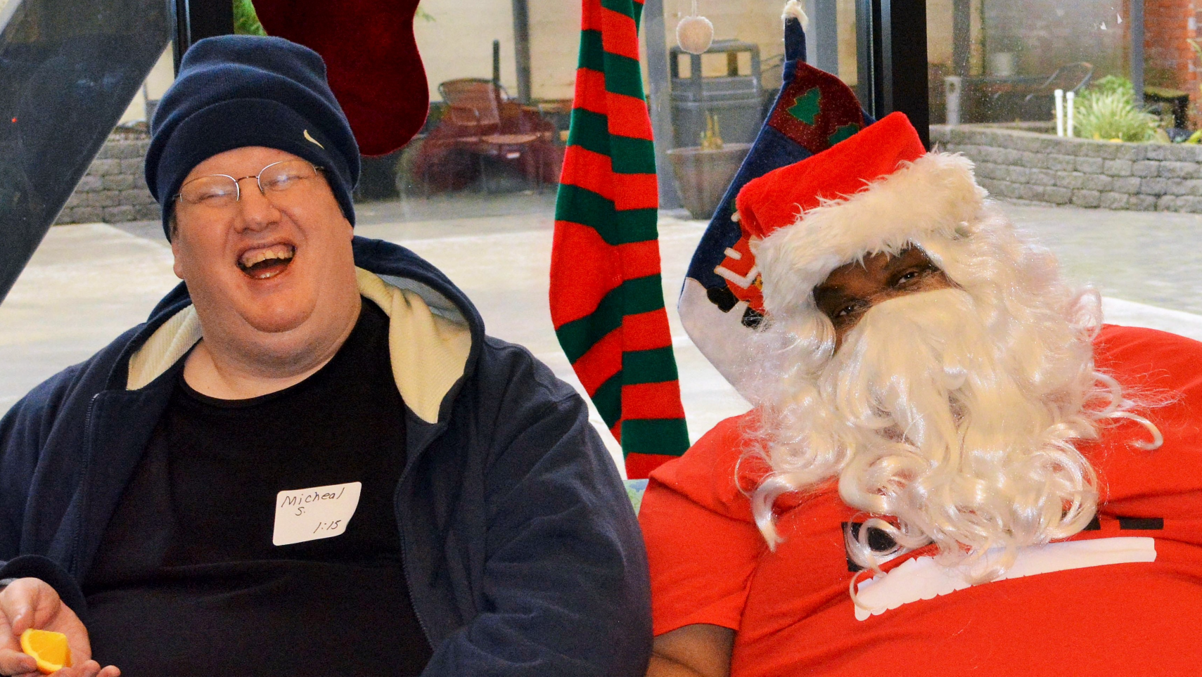 A middle age man with glasses and a beanie laughs next to santa