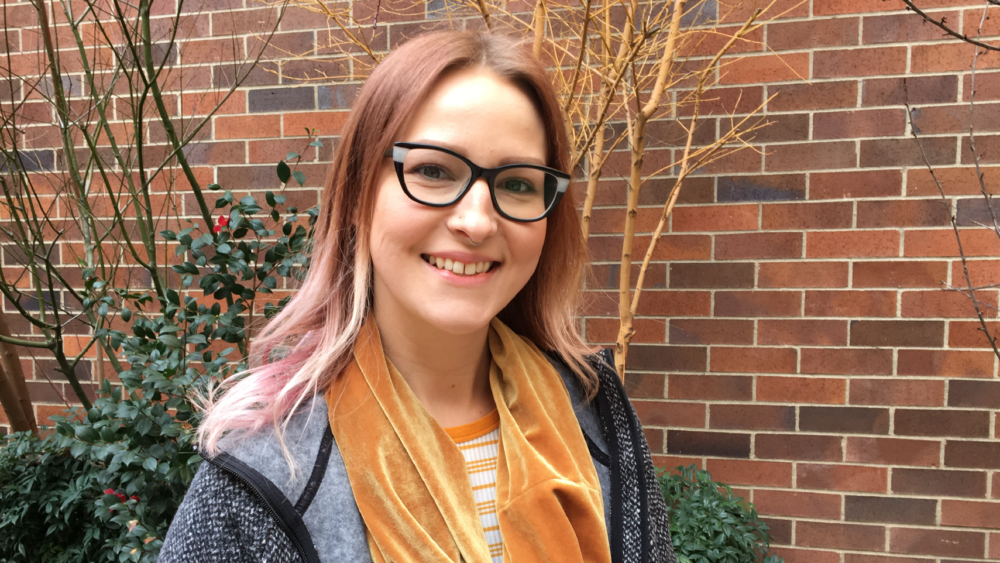 A young female volunteer looking towards you, standing in front of a brick wall.