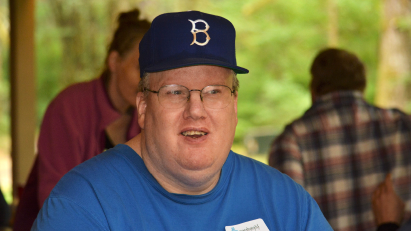 Mid-age male with a baseball hat and glasses looking at you, with nature and other people in the background