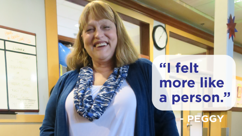 A smiling blonde woman in her 50's wearing a blue cardigan and using a walker inside a building