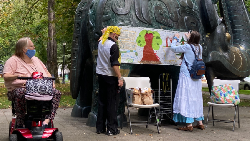 Two adults post a hand-colored poster on a statue outdoors and a person in a motorized scooter looks on