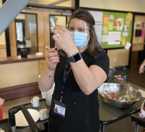 A nurse in protective gear inserts a syringe into a bottle of vaccine