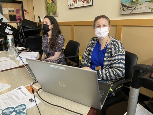 A female sits behind a table with a laptop, getting ready to check in people for the vaccine clinic