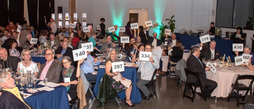 Interior shot of a room with at least five round tables visible, each seating several adults in business casual clothing. Many individuals are holding up white sheets of paper with black numbers on them and looking towards the camera.