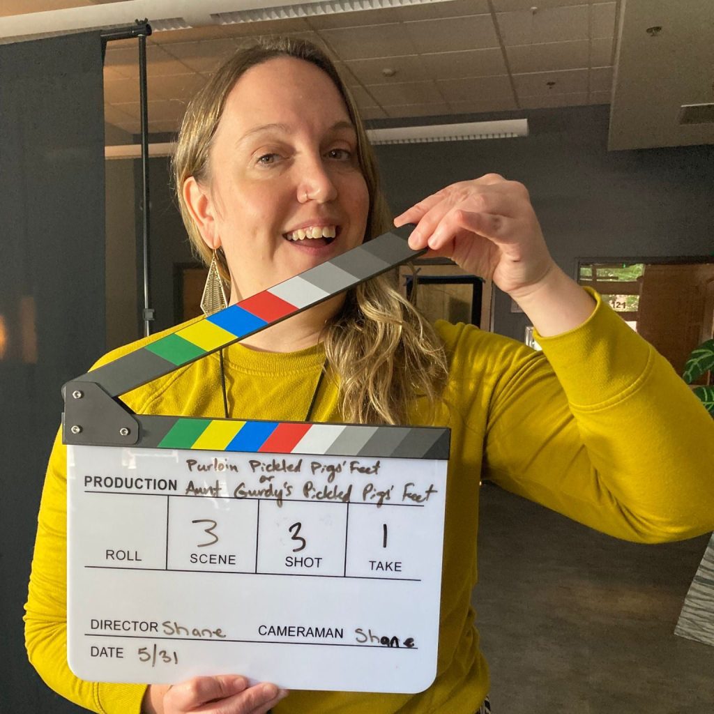 White woman smiling at camera wearing long sleeve yellow sweater and holding clapperboard up to show the scene and shot number of filming