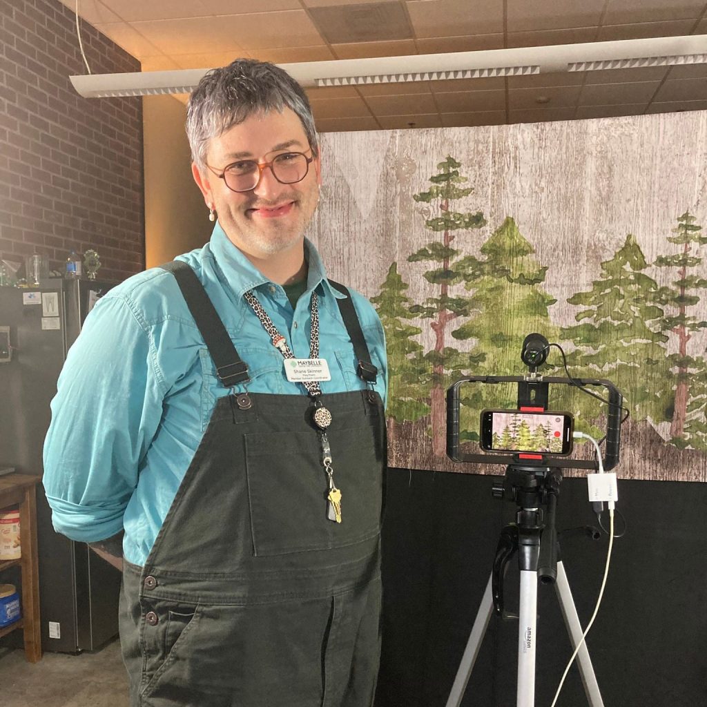 White man with glasses and blue long-sleeve shirt under dark green overalls smiling at the camera and standing next to a tripod, infront of a print background of green trees