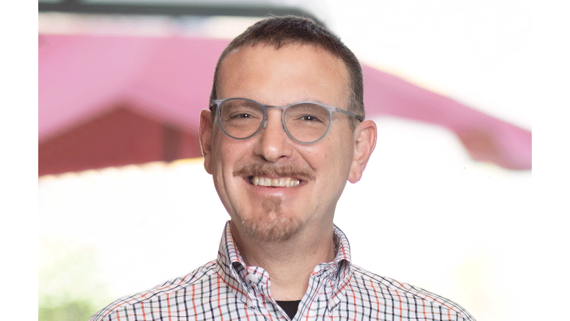 Middle aged white man with dark hair, glasses, a mustache, and goatee wearing a light colored plaid button-down collared shirt