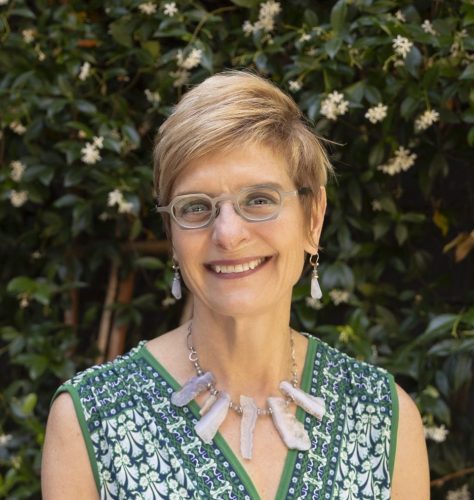 Middle aged white woman with blond, short hair, wearing glasses, earrings, and a large necklace, smiling at the camera outside infront of a wall of plants