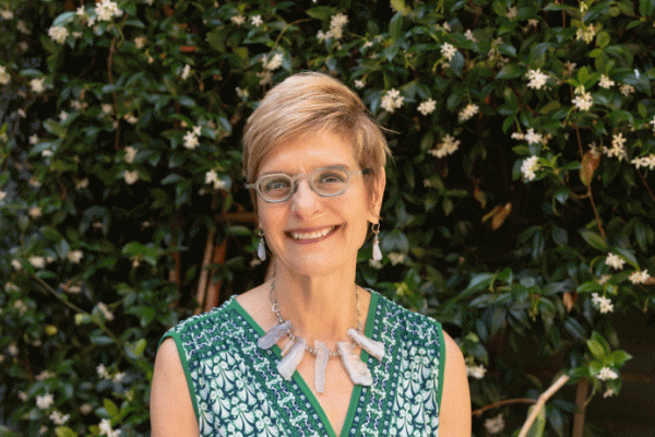 Middle aged white woman with blond, short hair, wearing glasses, earrings, and a large necklace, smiling at the camera outside infront of a wall of plants