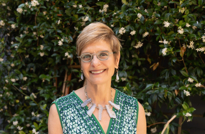 Middle aged white woman with blond, short hair, wearing glasses, earrings, and a large necklace, smiling at the camera outside infront of a wall of plants