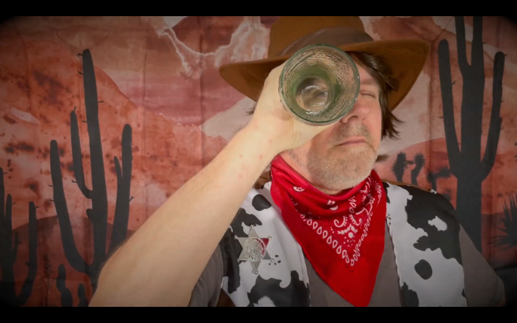 White man holding up glass vase or tube to eye, looking through it, while wearing red bandana, cow-print vest, and cowboy hat infront of painted red backdrop of dessert scene with two cacti