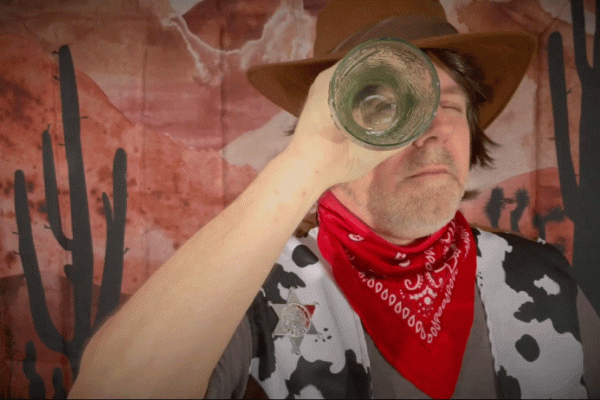 White man holding up glass vase or tube to eye, looking through it, while wearing red bandana, cow-print vest, and cowboy hat infront of painted red backdrop of dessert scene with two cacti