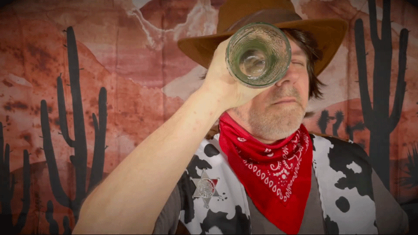 White man holding up glass vase or tube to eye, looking through it, while wearing red bandana, cow-print vest, and cowboy hat infront of painted red backdrop of dessert scene with two cacti