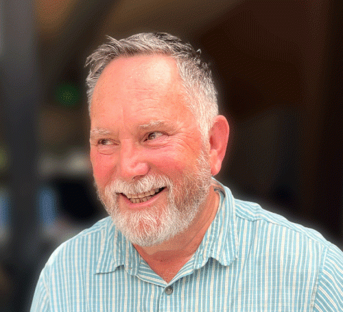 Older white man with white and black hair, white mustache and close-cropped beard, wearing a blue and white striped shirt, smiling and looking away from the camera with a dark background