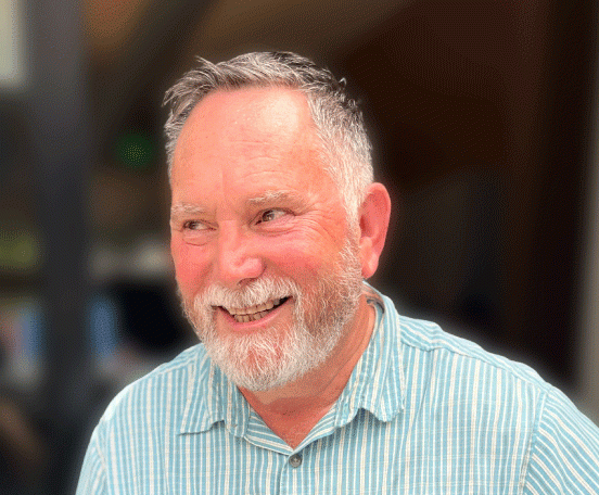 Older white man with white and black hair, white mustache and close-cropped beard, wearing a blue and white striped shirt, smiling and looking away from the camera with a dark background