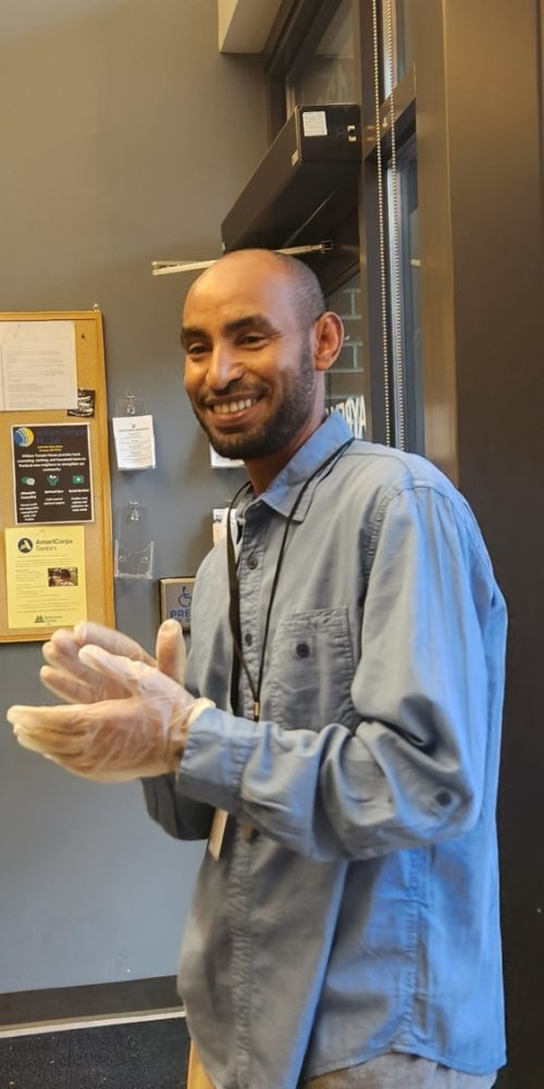 A middle-aged, balding man with darker skin looking at the camera inside a building in front of a window