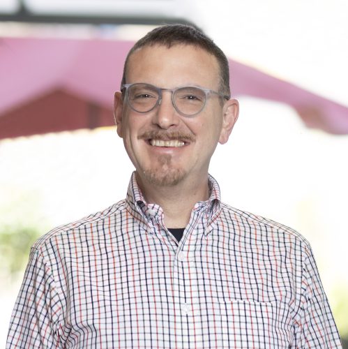 Middle aged white man with dark hair, glasses, a mustache, and goatee wearing a light colored plaid button-down collared shirt