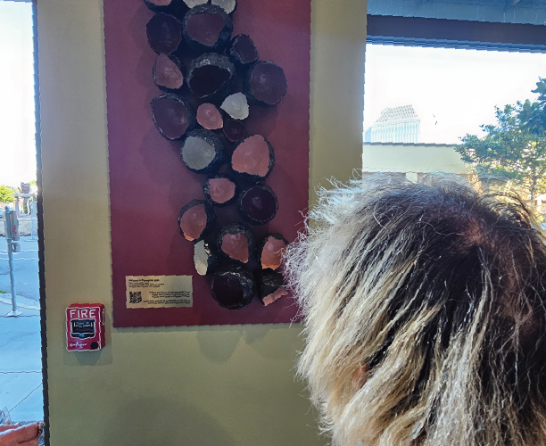 The back of a person's head looking up at an artwork hanging inside on a pillar. The artwork is a large red background, covered in different colored paper mache cups, black on the outside with their colorful interiors facing out towards the viewer