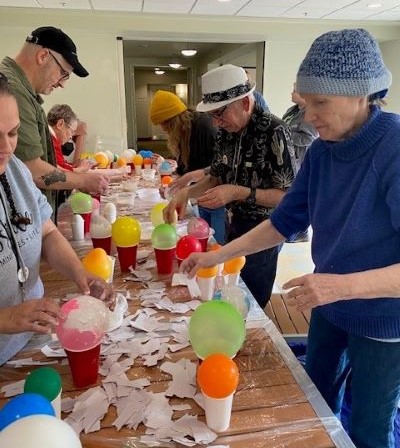 Several people stand on both sides of a long table, where colorful balloons sit on top of cups, as the individuals attach paper to the balloons, making paper mache shapes