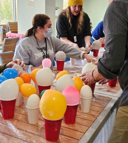 White, yellow, and pink balloons sit on top of red cups atop a table, while people stand and sit around the table. A person in a grey shirt and with a mask on, reaches across the table while a person standing at the end of the table in a black shirt and yellow beanie gives directions