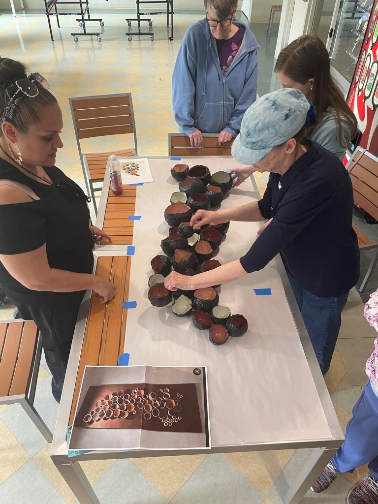 Individuals stand around a table covered in white paper, with many paper mache cups on top. The paper mache cups are black on the outside with colorful interiors, and the individuals standing around are touching them, laying them out on the white paper