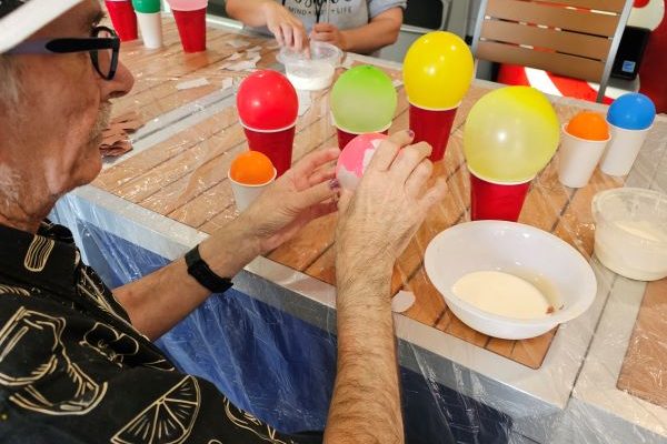 yellow, green, and pink balloons sit on top of red plastic cups, atop a table next to a small bowl of white glue, while an older persons' hands work on pasting paper onto the balloon in front of them