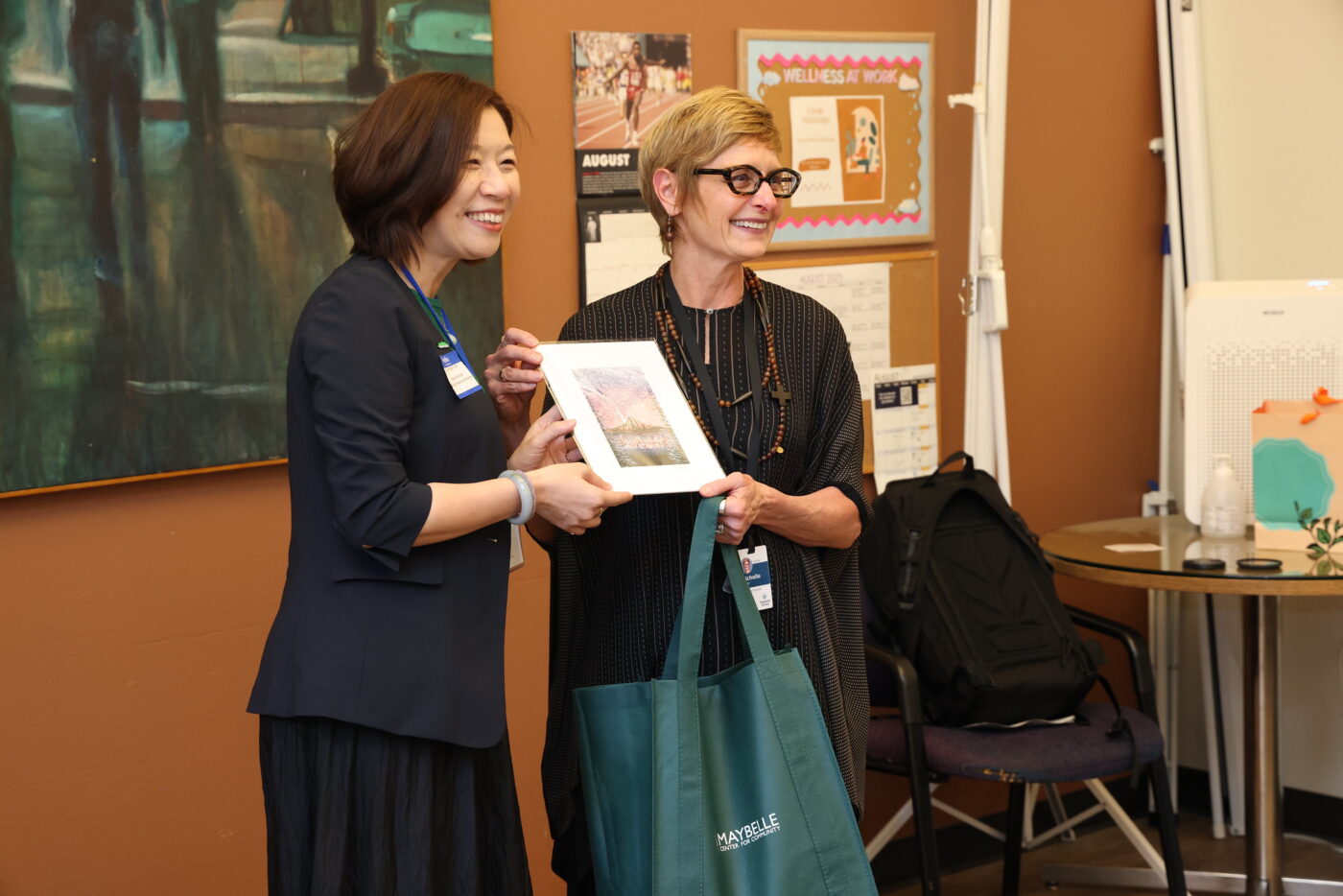 Two women with short hair stand smiling at the front of a room, both holding forward a print.