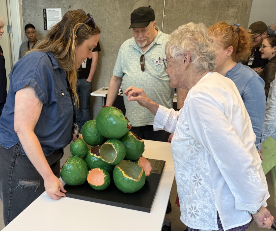 Four people standing around a table, looking down at a small bright-green bulbous sculpture being placed down on the table by one of the four people