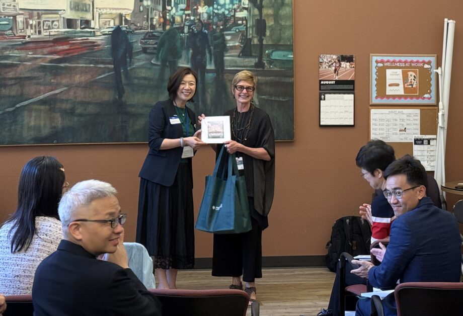 Two women with short hair stand smiling at the front of a room, both holding forward a print to show the room of people seated in front of them.