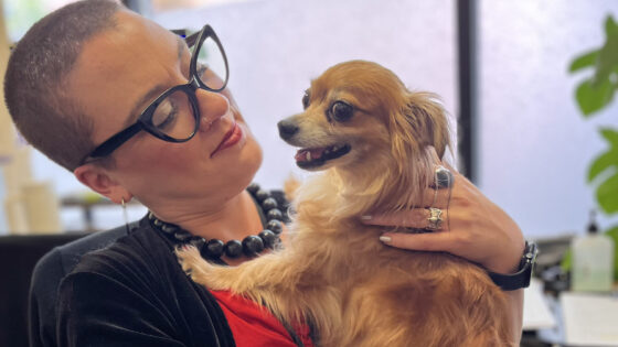 A white presenting woman with dark glasses and buzz-cut hair sits holding a small dog and looking admiringly at the dog who is resting on one of her legs while their front paws are on her chest, their closes almost face to face