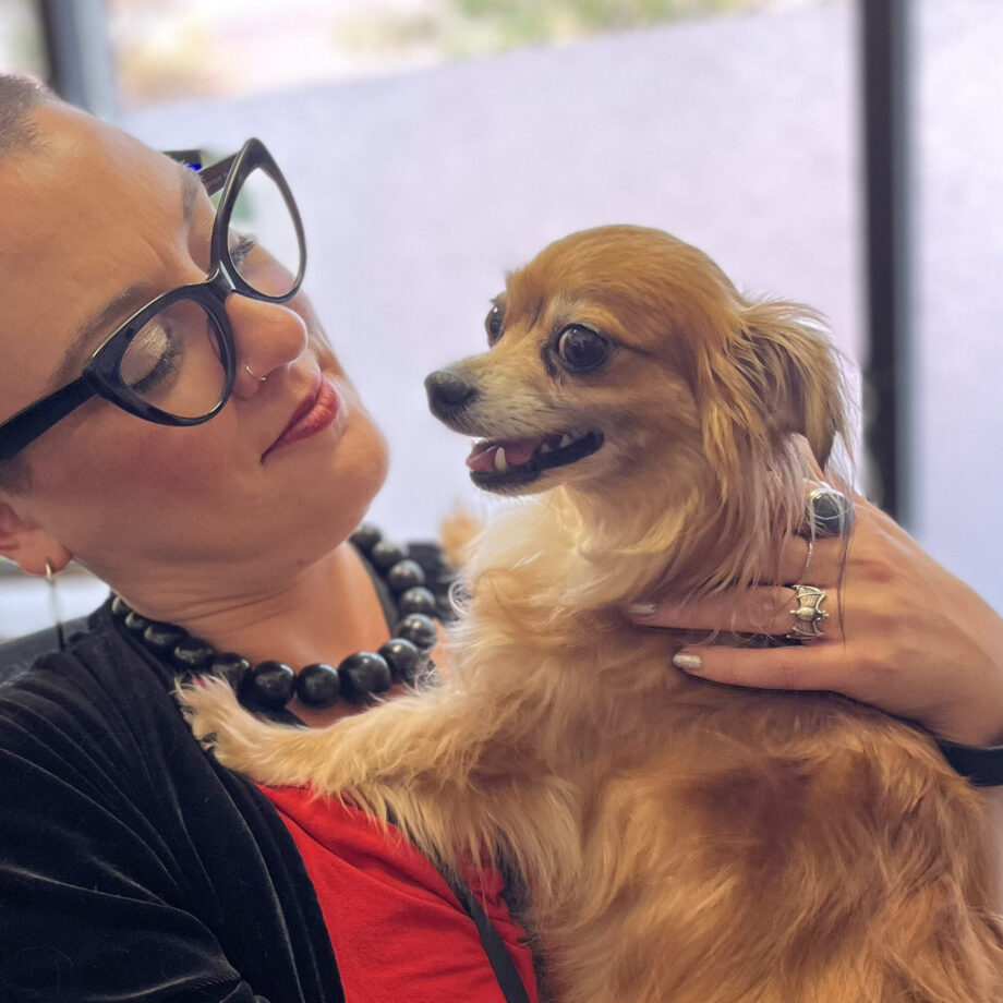 A white presenting woman with dark glasses and buzz-cut hair sits holding a small dog and looking admiringly at the dog who is resting on one of her legs while their front paws are on her chest, their closes almost face to face