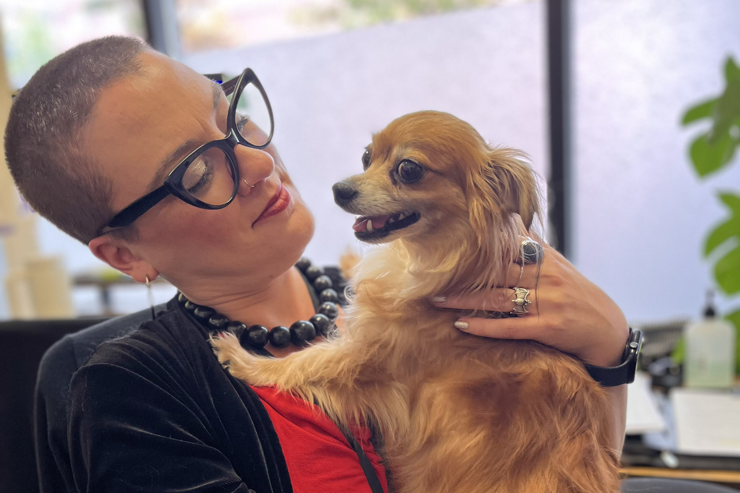 A white presenting woman with dark glasses and buzz-cut hair sits holding a small dog and looking admiringly at the dog who is resting on one of her legs while their front paws are on her chest, their closes almost face to face