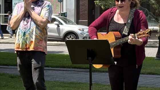 Two adults stand in the grass singing, on the left a white presenting man has black pants and tye-dye t-shirt, clapping his hands together. On the right, a white presenting woman stands, playing a guitar and looking at a music stand in front of her.
