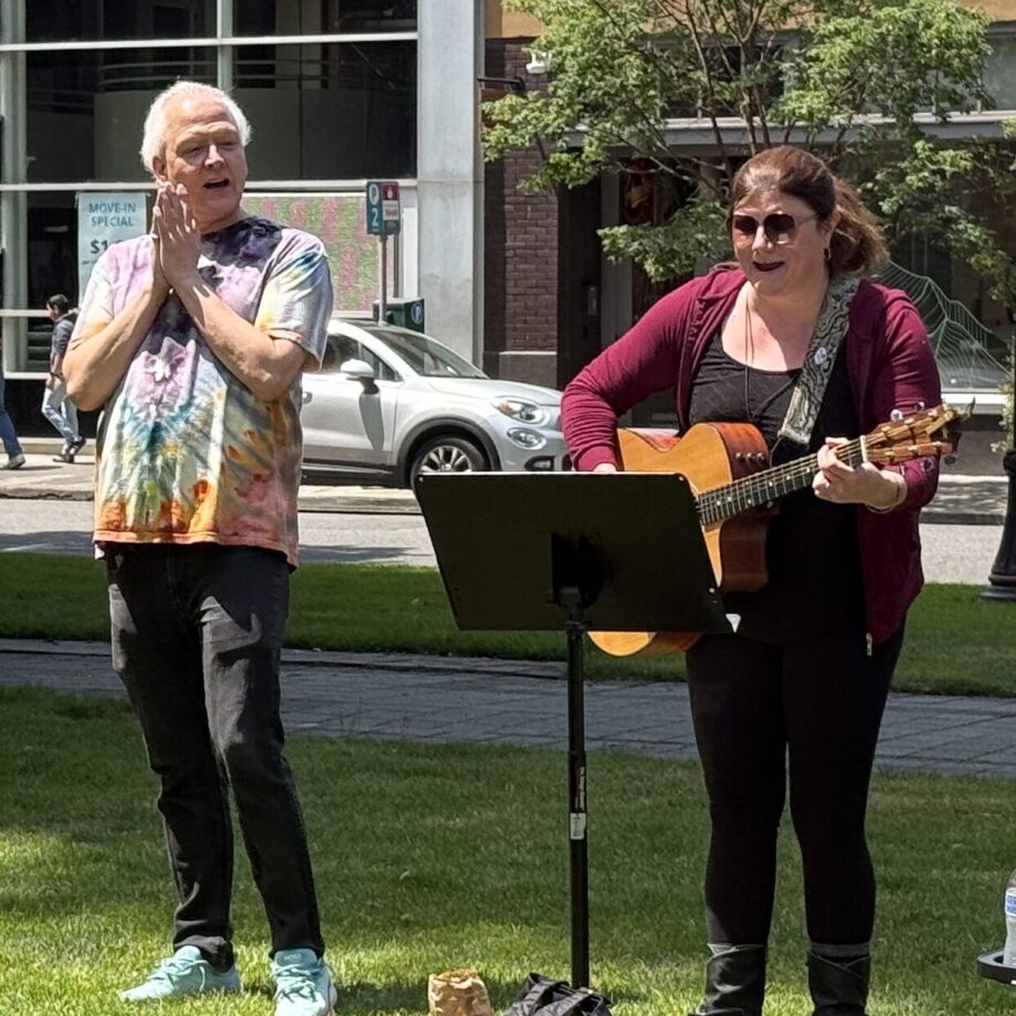 Two adults stand in the grass singing, on the left a white presenting man has black pants and tye-dye t-shirt, clapping his hands together. On the right, a white presenting woman stands, playing a guitar and looking at a music stand in front of her.