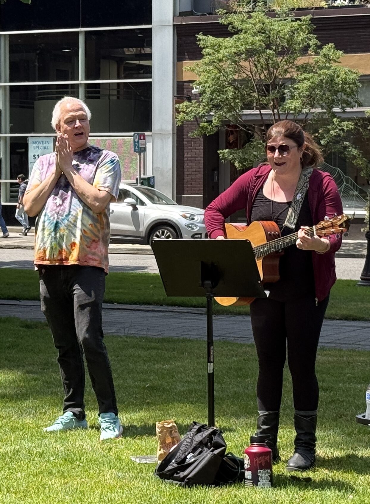 Two adults stand in the grass singing, on the left a white presenting man has black pants and tye-dye t-shirt, clapping his hands together. On the right, a white presenting woman stands, playing a guitar and looking at a music stand in front of her.