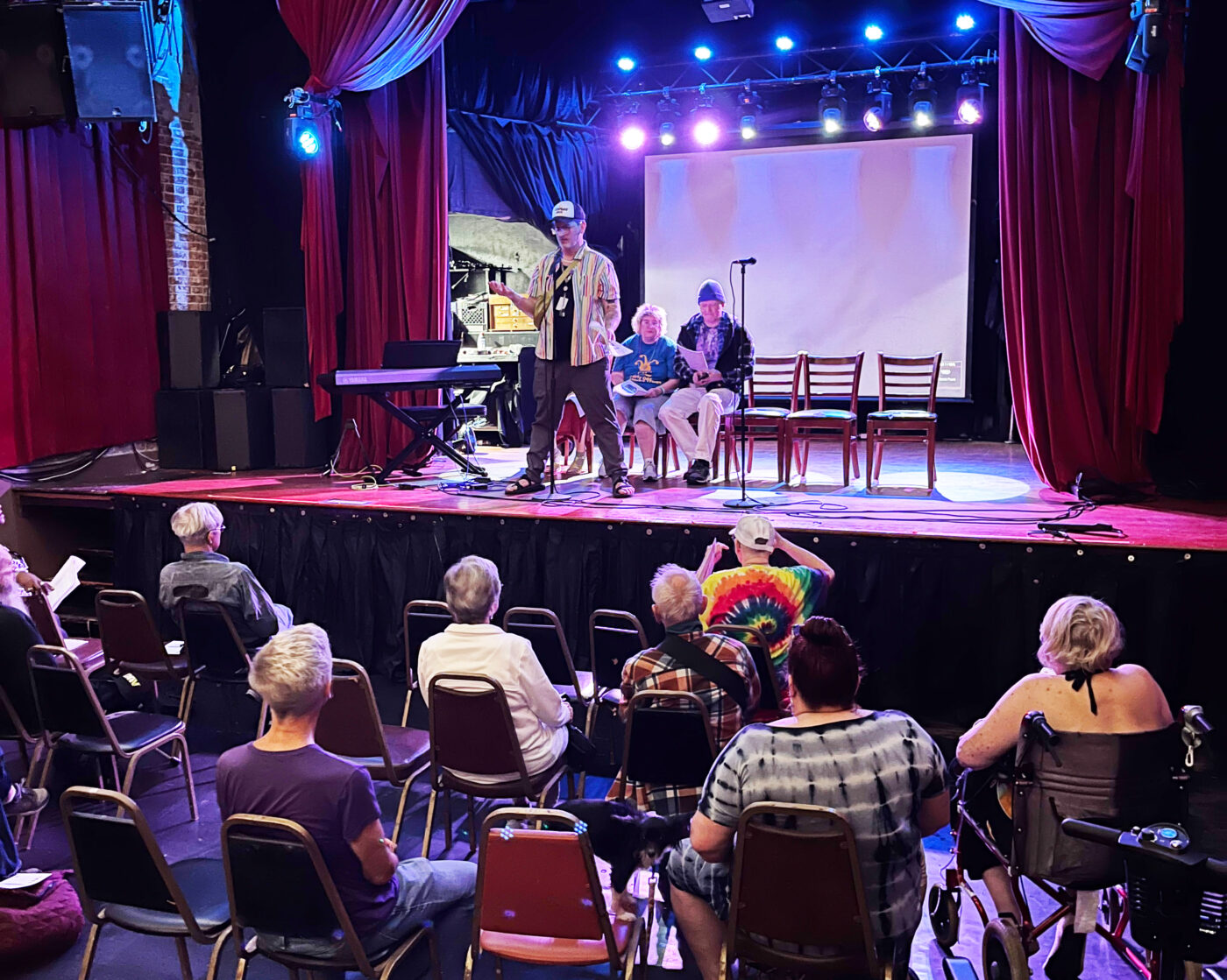 An individual stands at the front center of a performance stage, with two people seated in chairs behind them on stage. A mic on a stand and a keyboard are also on stage with these individuals. In front and below the stage are individual seats and the back of several people's heads are visible as they watch the central, standing figure talk on stage.